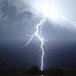 Bright lightning bolts strike downward over a dark, stormy sky above a silhouetted landscape at dusk/dark.