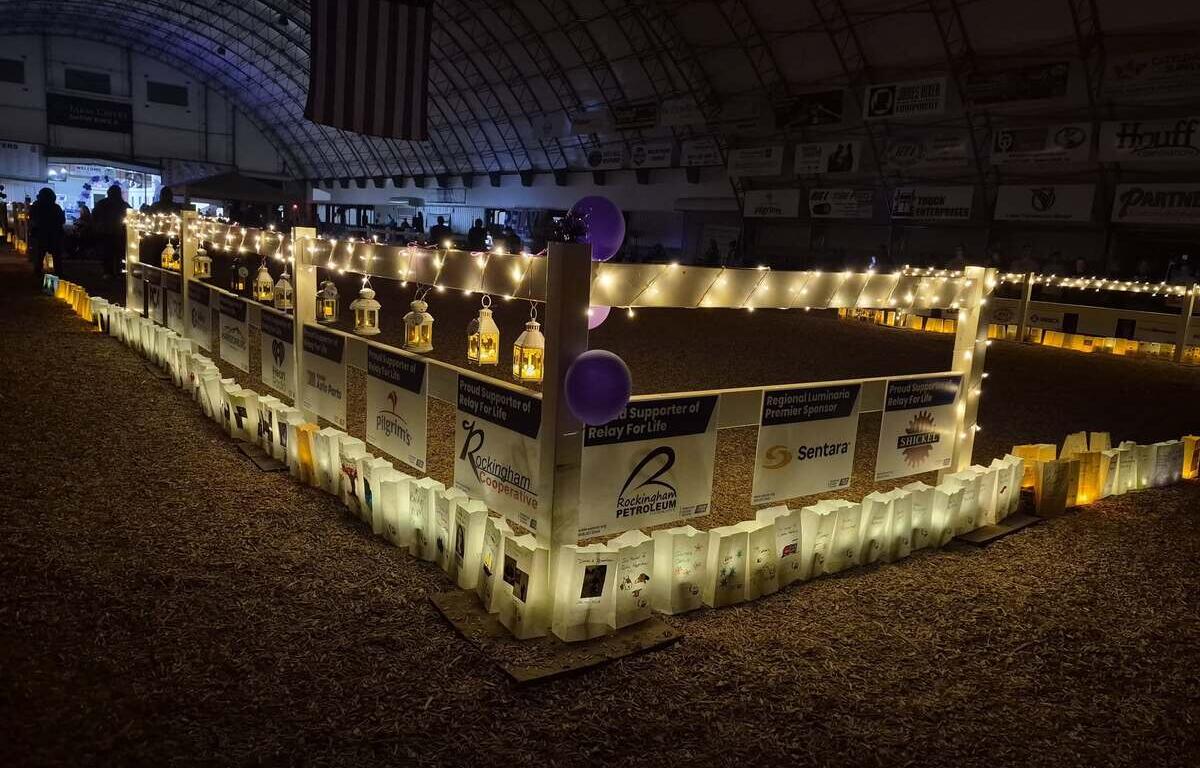 A festive indoor event in a dim arena with a lit perimeter fence decorated with string lights, lanterns, posters, and purple balloons for Relay for Life.