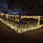 A festive indoor event in a dim arena with a lit perimeter fence decorated with string lights, lanterns, posters, and purple balloons for Relay for Life.