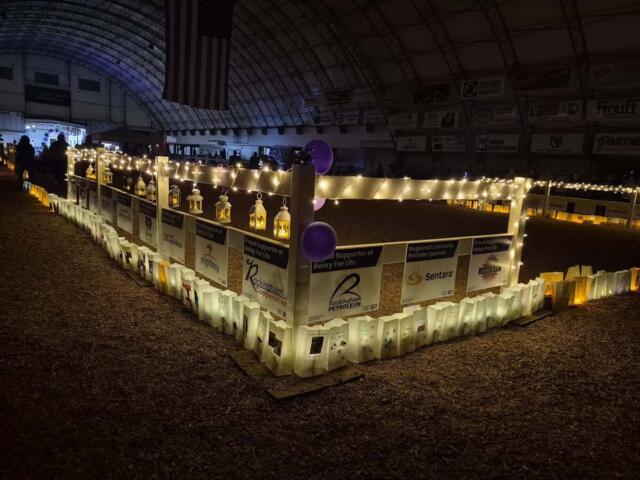 A festive indoor event in a dim arena with a lit perimeter fence decorated with string lights, lanterns, posters, and purple balloons for Relay for Life.