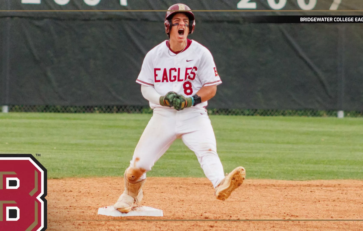 Baseball player in a white Eagles uniform slides into first base on the dirt infield, with a large team logo in the foreground and a green outfield behind him.