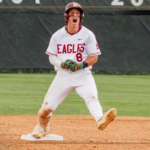 Baseball player in a white Eagles uniform slides into first base on the dirt infield, with a large team logo in the foreground and a green outfield behind him.