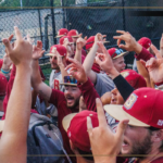 Bridgewater College Eagles baseball players celebrate with raised hands in a huddle on the field, a large B logo visible on the left.