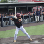 Baseball batter in black swings at home plate as teammates watch from the dugout; Bridgewater College Eagles insignia bottom-left.