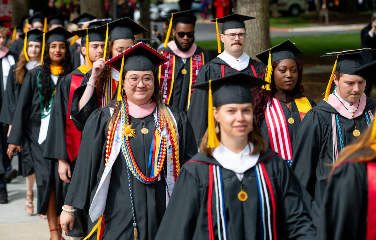 Group of graduates in black cap-and-gown procession outdoors, front center student wearing colorful cords and glasses.