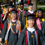 Group of graduates in black cap-and-gown procession outdoors, front center student wearing colorful cords and glasses.