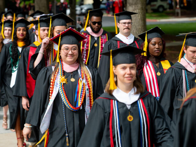Group of graduates in black cap-and-gown procession outdoors, front center student wearing colorful cords and glasses.