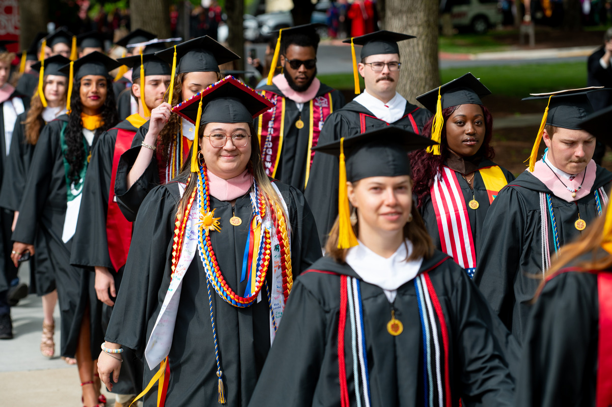 Group of graduates in black cap-and-gown procession outdoors, front center student wearing colorful cords and glasses.