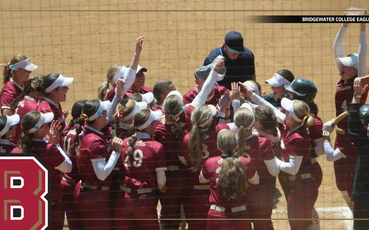 Bridgewater College Eagles softball players in maroon uniforms celebrate in a huddle on the dirt field, coach at the center.