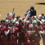 Bridgewater College Eagles softball players in maroon uniforms celebrate in a huddle on the dirt field, coach at the center.
