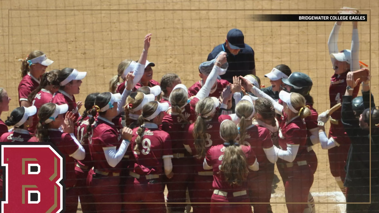 Bridgewater College Eagles softball players in maroon uniforms celebrate in a huddle on the dirt field, coach at the center.