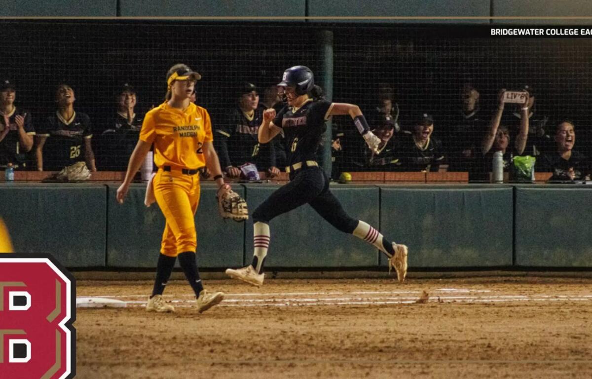 Softball scene at night: a runner in a dark uniform slides toward home plate while a yellow-clad fielder watches nearby, with cheering fans behind the fence and a large red team logo in the foreground.