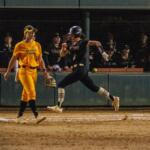 Softball scene at night: a runner in a dark uniform slides toward home plate while a yellow-clad fielder watches nearby, with cheering fans behind the fence and a large red team logo in the foreground.