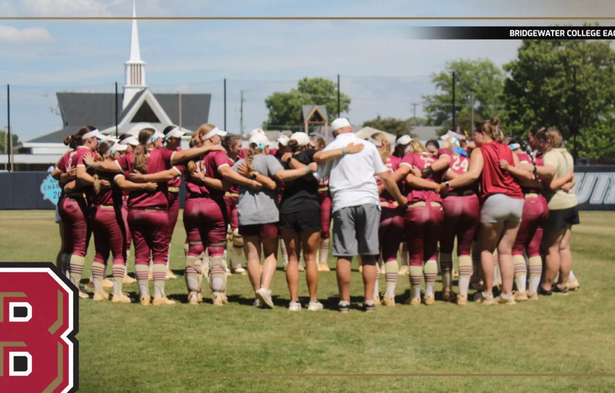 Bridgewater College Eagles softball team huddles in a circle on the field before a game, maroon uniforms visible.