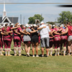 Bridgewater College Eagles softball team huddles in a circle on the field before a game, maroon uniforms visible.