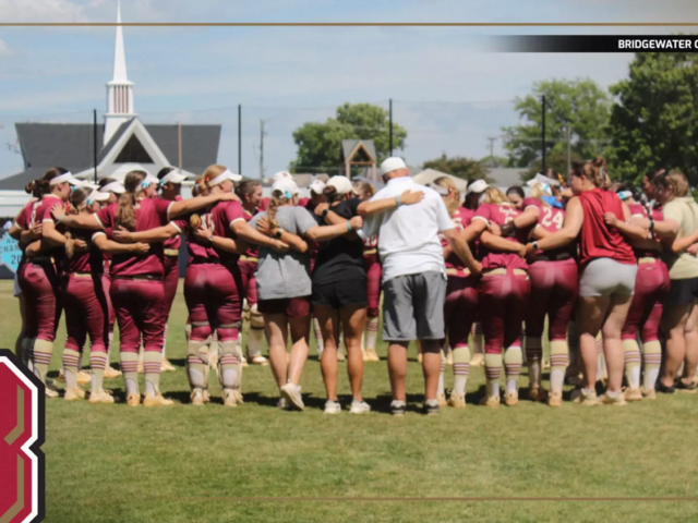 Bridgewater College Eagles softball team huddles in a circle on the field before a game, maroon uniforms visible.