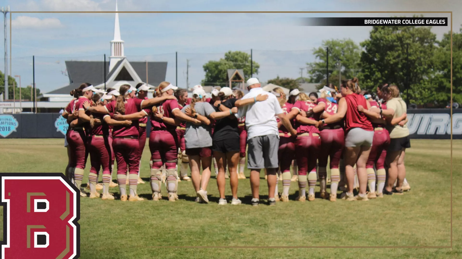 Bridgewater College Eagles softball team huddles in a circle on the field before a game, maroon uniforms visible.