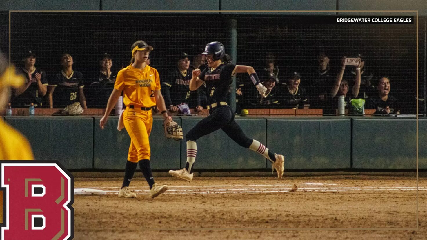Softball scene at night: a runner in a dark uniform slides toward home plate while a yellow-clad fielder watches nearby, with cheering fans behind the fence and a large red team logo in the foreground.