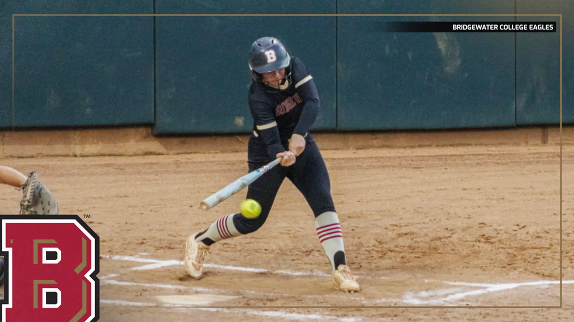 Female softball batter in a navy uniform swings at a pitched ball at home plate, with a green outfield wall in the background and a large B logo foregrounded on the left.