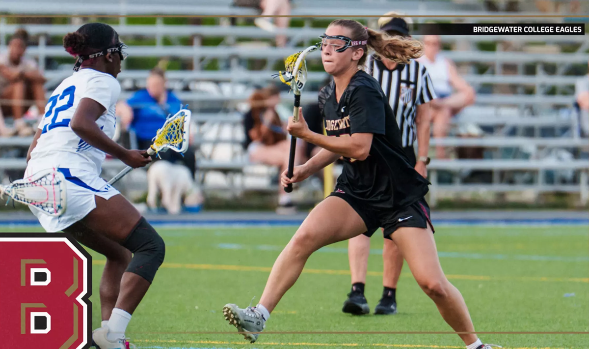 Lacrosse players in black and white uniforms compete for the ball on a turf field during a game, with a Bridgewater College Eagles logo visible in the corner.