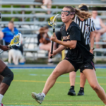 Lacrosse players in black and white uniforms compete for the ball on a turf field during a game, with a Bridgewater College Eagles logo visible in the corner.