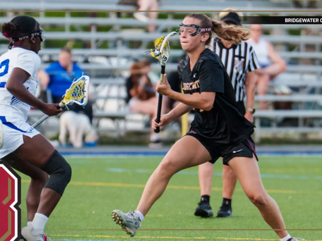 Lacrosse players in black and white uniforms compete for the ball on a turf field during a game, with a Bridgewater College Eagles logo visible in the corner.