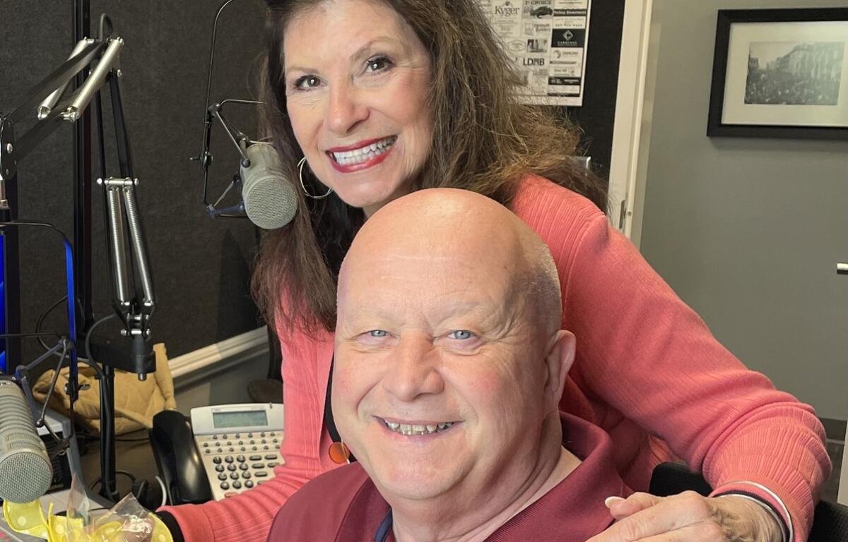 Smiling woman and man in a radio studio with microphones, a mixing board, and a gift basket on the desk in front of them.