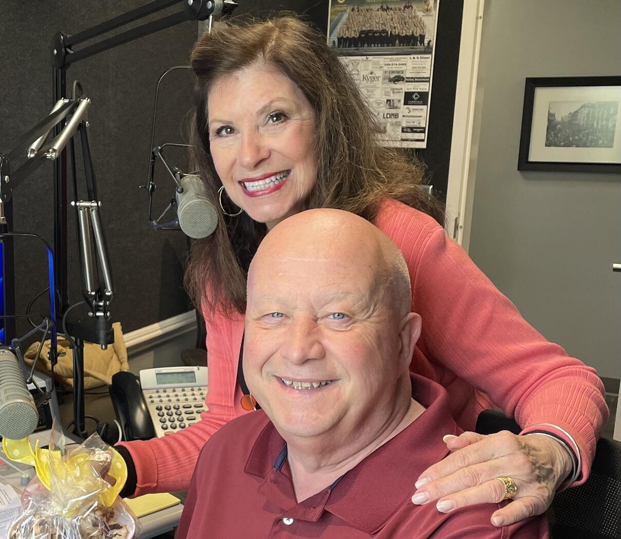 Smiling woman and man in a radio studio with microphones, a mixing board, and a gift basket on the desk in front of them.