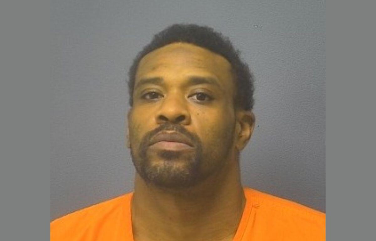 Mugshot portrait of a man with short dark hair and facial hair wearing an orange jail jumpsuit against a gray backdrop, looking at the camera.