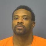 Mugshot portrait of a man with short dark hair and facial hair wearing an orange jail jumpsuit against a gray backdrop, looking at the camera.