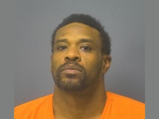 Mugshot portrait of a man with short dark hair and facial hair wearing an orange jail jumpsuit against a gray backdrop, looking at the camera.
