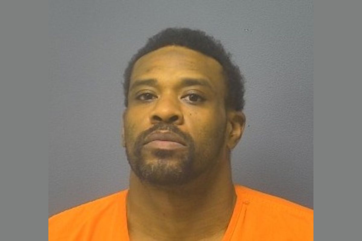 Mugshot portrait of a man with short dark hair and facial hair wearing an orange jail jumpsuit against a gray backdrop, looking at the camera.
