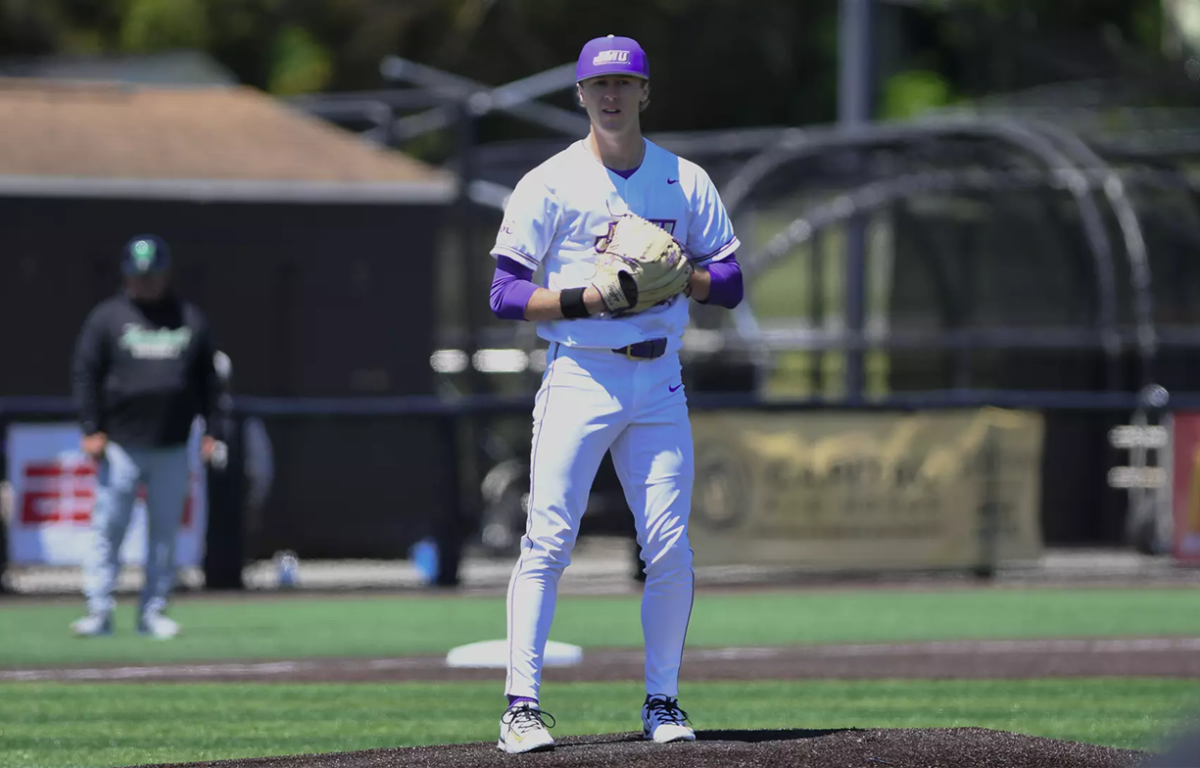 Baseball pitcher on the mound in a white and purple uniform, glove up and ready to pitch.