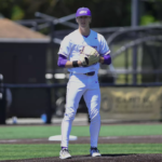 Baseball pitcher on the mound in a white and purple uniform, glove up and ready to pitch.