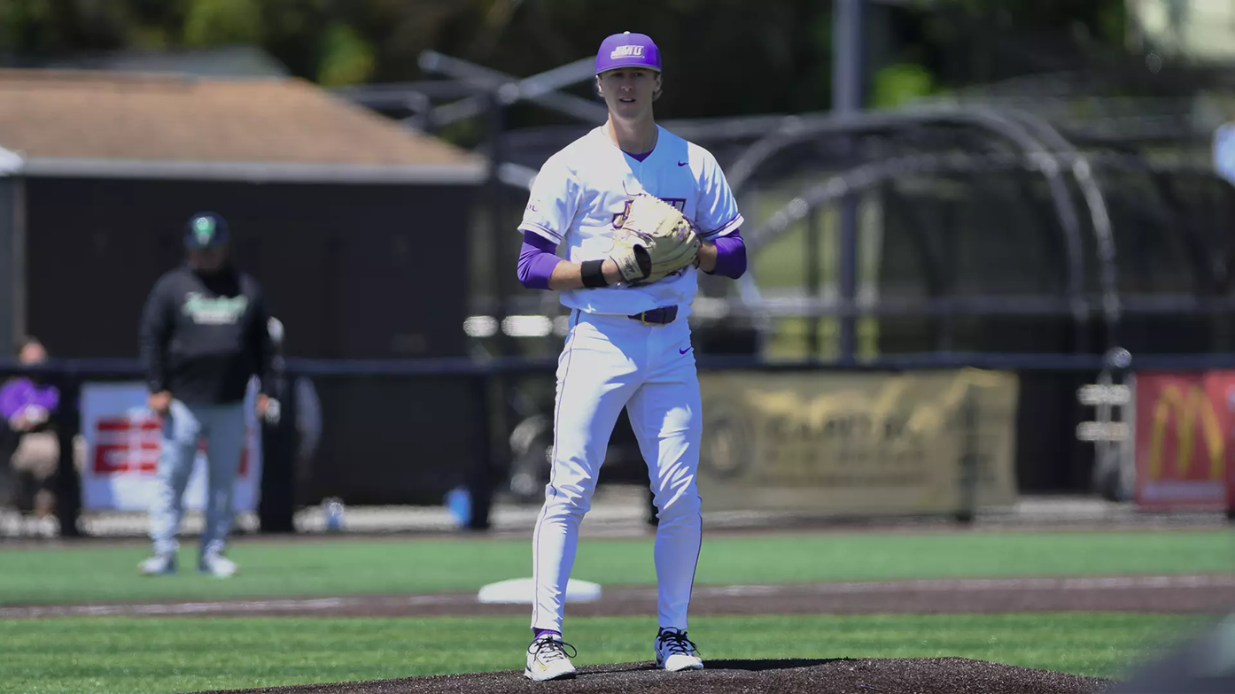 Baseball pitcher on the mound in a white and purple uniform, glove up and ready to pitch.