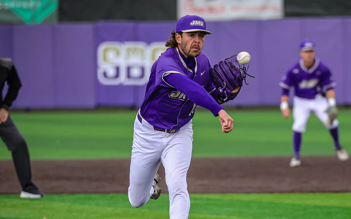 Baseball pitcher in a purple uniform winds up a pitch on a field with a purple outfield wall behind him.