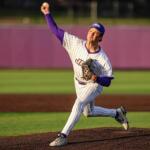 Baseball pitcher in a pinstriped uniform delivering a throw on the mound, purple accents, purple cap, with a fielder in the background