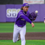 Baseball pitcher in a purple uniform winds up a pitch on a field with a purple outfield wall behind him.
