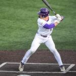 Baseball batter in white uniform swinging at a pitch, catcher in yellow crouched behind home plate.