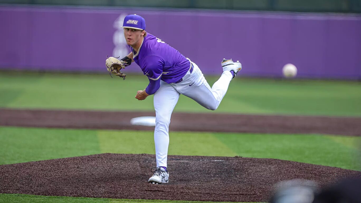 College baseball pitcher in purple uniform throwing from the mound, ball in midair behind him.