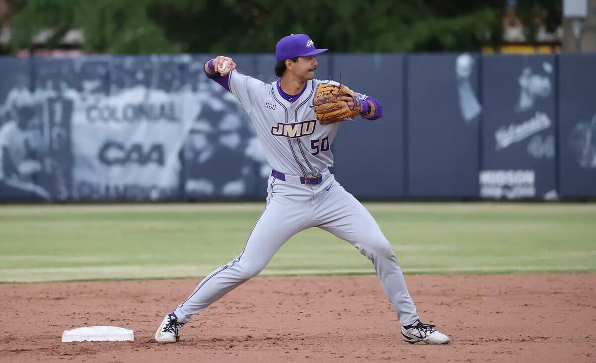 Baseball pitcher in gray and purple JMU uniform delivering a pitch on the dirt mound with a blue outfield wall behind him.