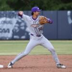 Baseball pitcher in gray and purple JMU uniform delivering a pitch on the dirt mound with a blue outfield wall behind him.