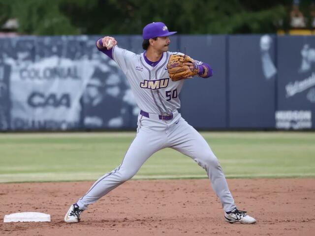 Baseball pitcher in gray and purple JMU uniform delivering a pitch on the dirt mound with a blue outfield wall behind him.