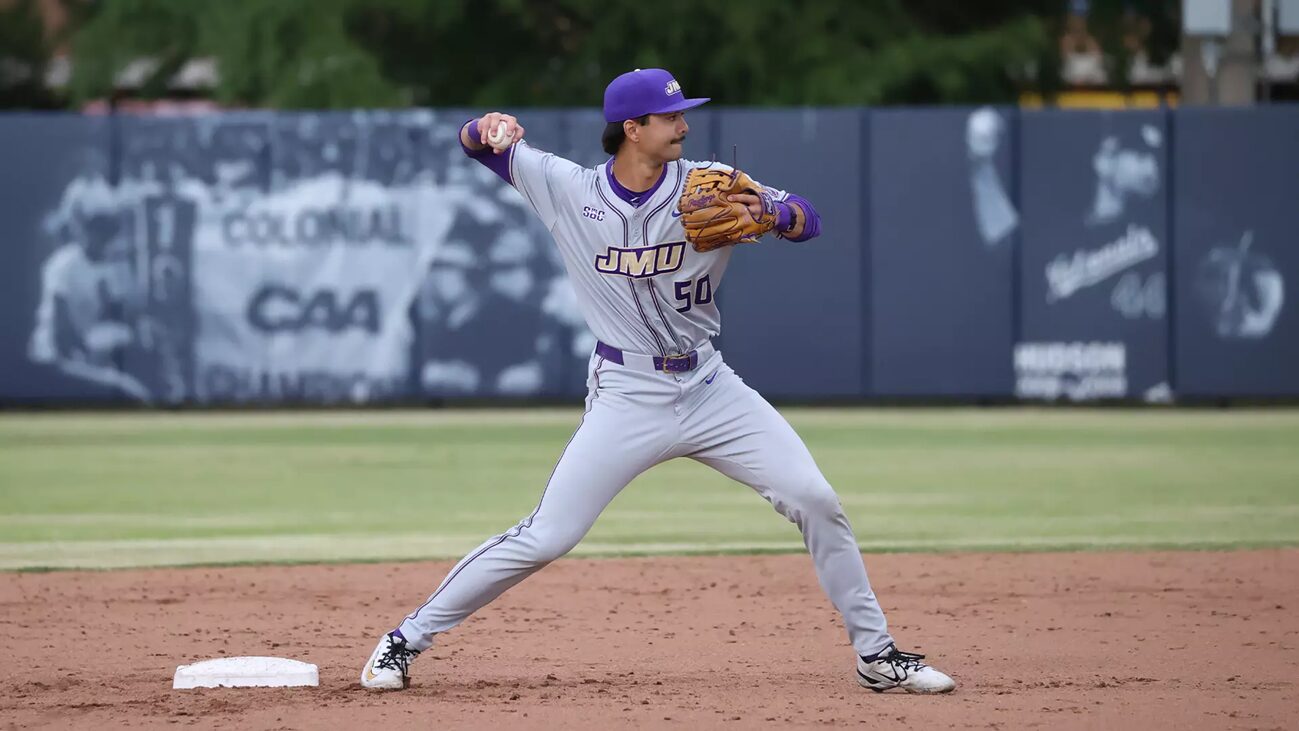 Baseball pitcher in gray and purple JMU uniform delivering a pitch on the dirt mound with a blue outfield wall behind him.