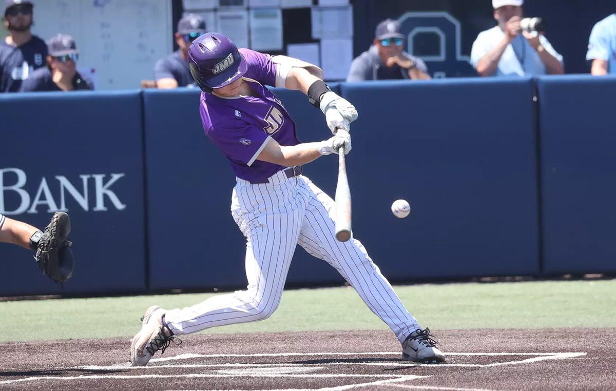Baseball batter in a purple uniform swings at a pitch on a dirt batter's box; ball near the bat with a catcher visible to the left and a blue padded wall in the background.