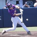 Baseball batter in a purple uniform swings at a pitch on a dirt batter's box; ball near the bat with a catcher visible to the left and a blue padded wall in the background.