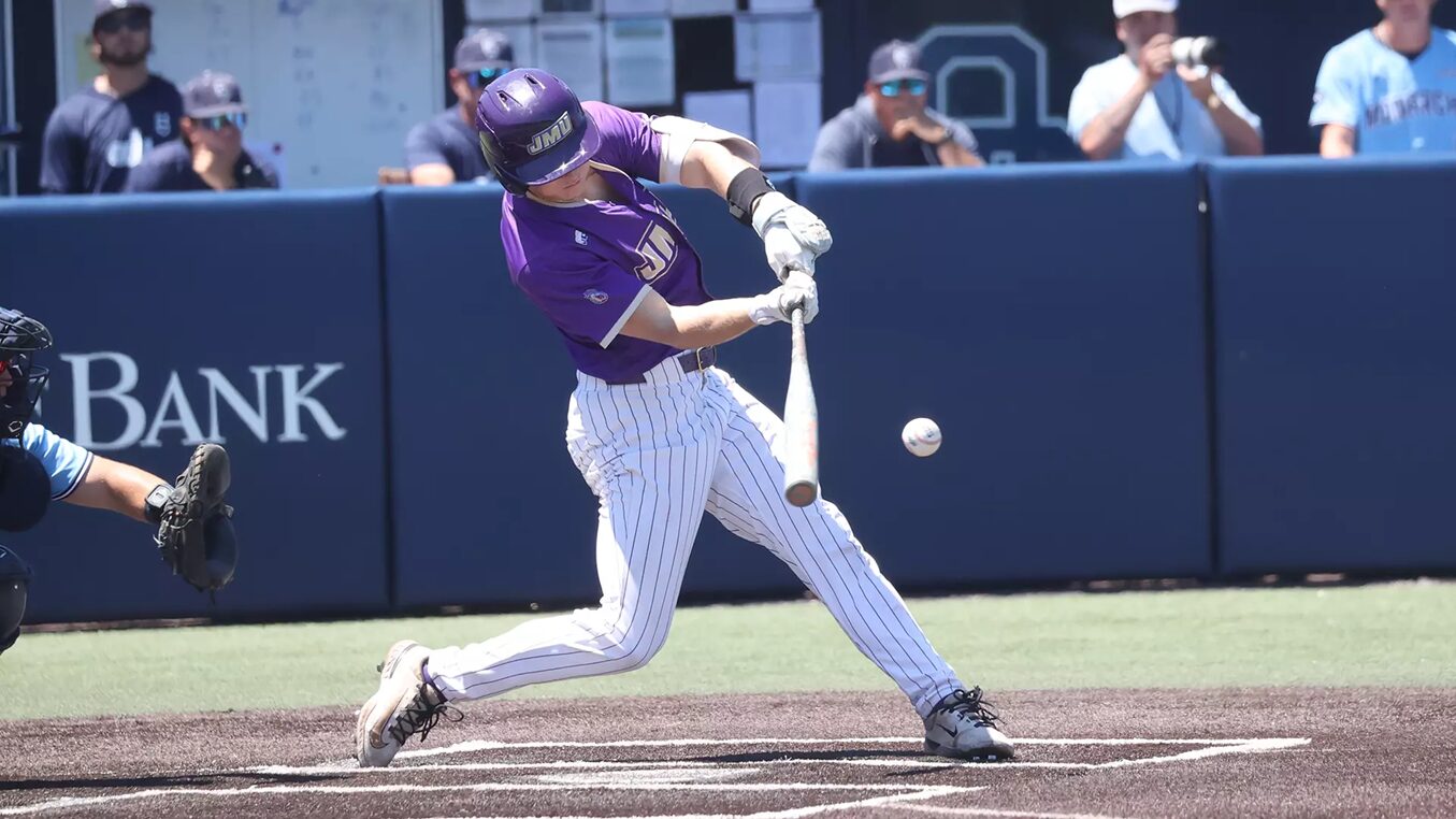 Baseball batter in a purple uniform swings at a pitch on a dirt batter's box; ball near the bat with a catcher visible to the left and a blue padded wall in the background.