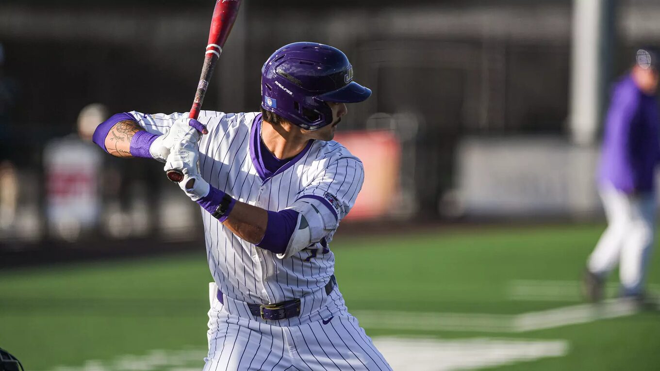 Baseball player in a white pinstripe uniform swings a bat, wearing a purple helmet and sleeves on a green field.