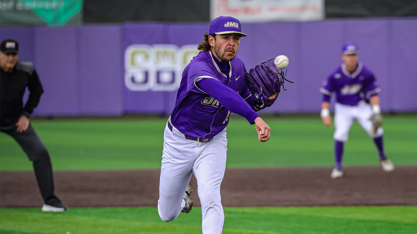 Baseball pitcher in a purple uniform winds up a pitch on a field with a purple outfield wall behind him.