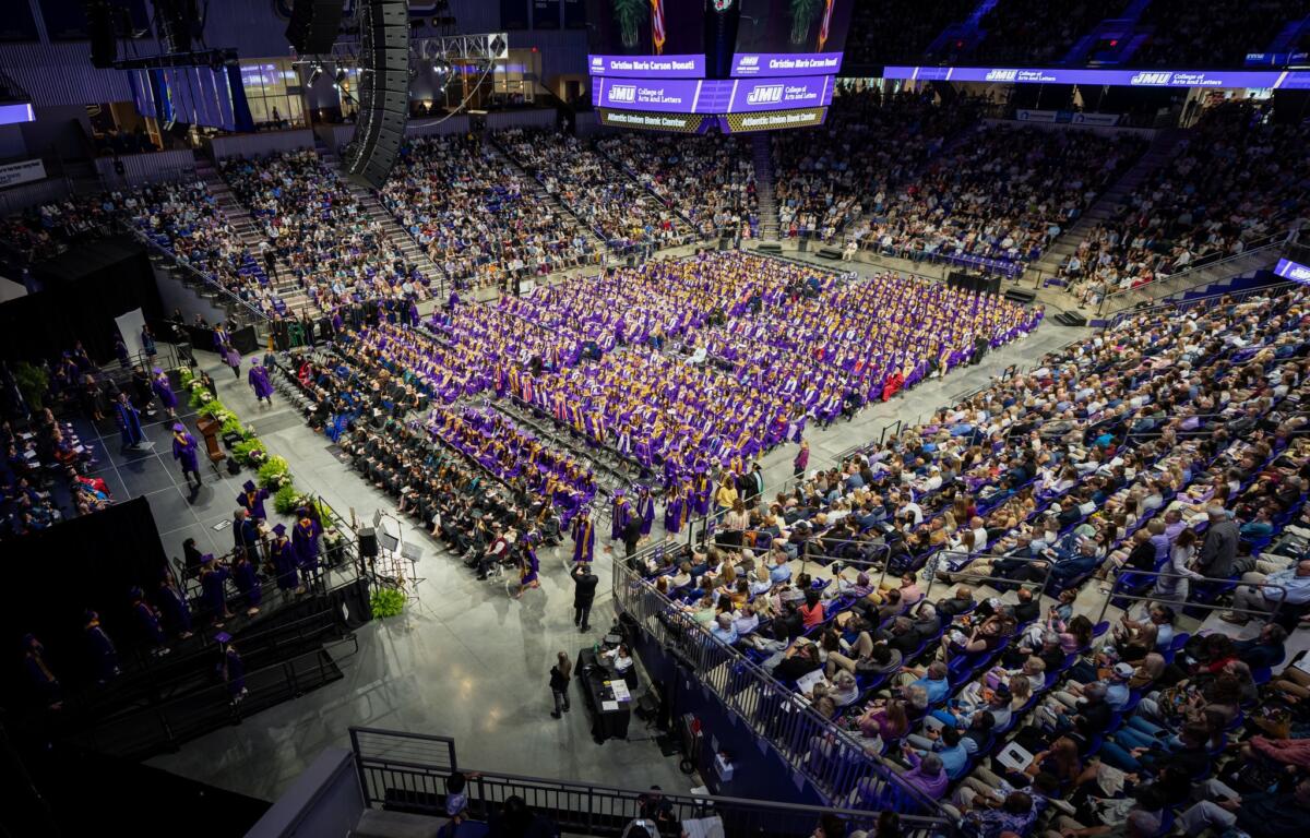 Wide view of a college graduation ceremony in an arena; hundreds of graduates in purple gowns sit in rows with a large audience surrounding them, stage on left.
